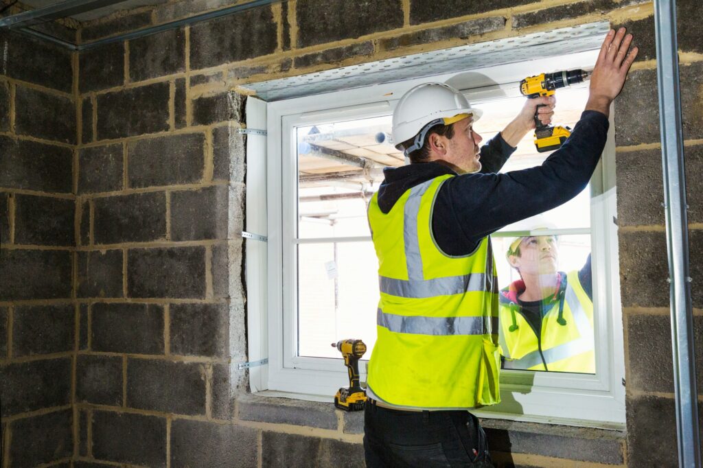 A workman on a construction site, a builder in hard hat using an electric drill on a window sill.