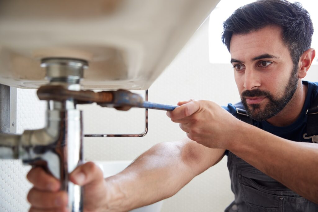 Male Plumber Using Wrench To Fix Leaking Sink In Home Bathroom