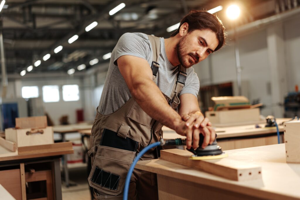 Young carpenter sanding wood piece in workshop in furniture factory